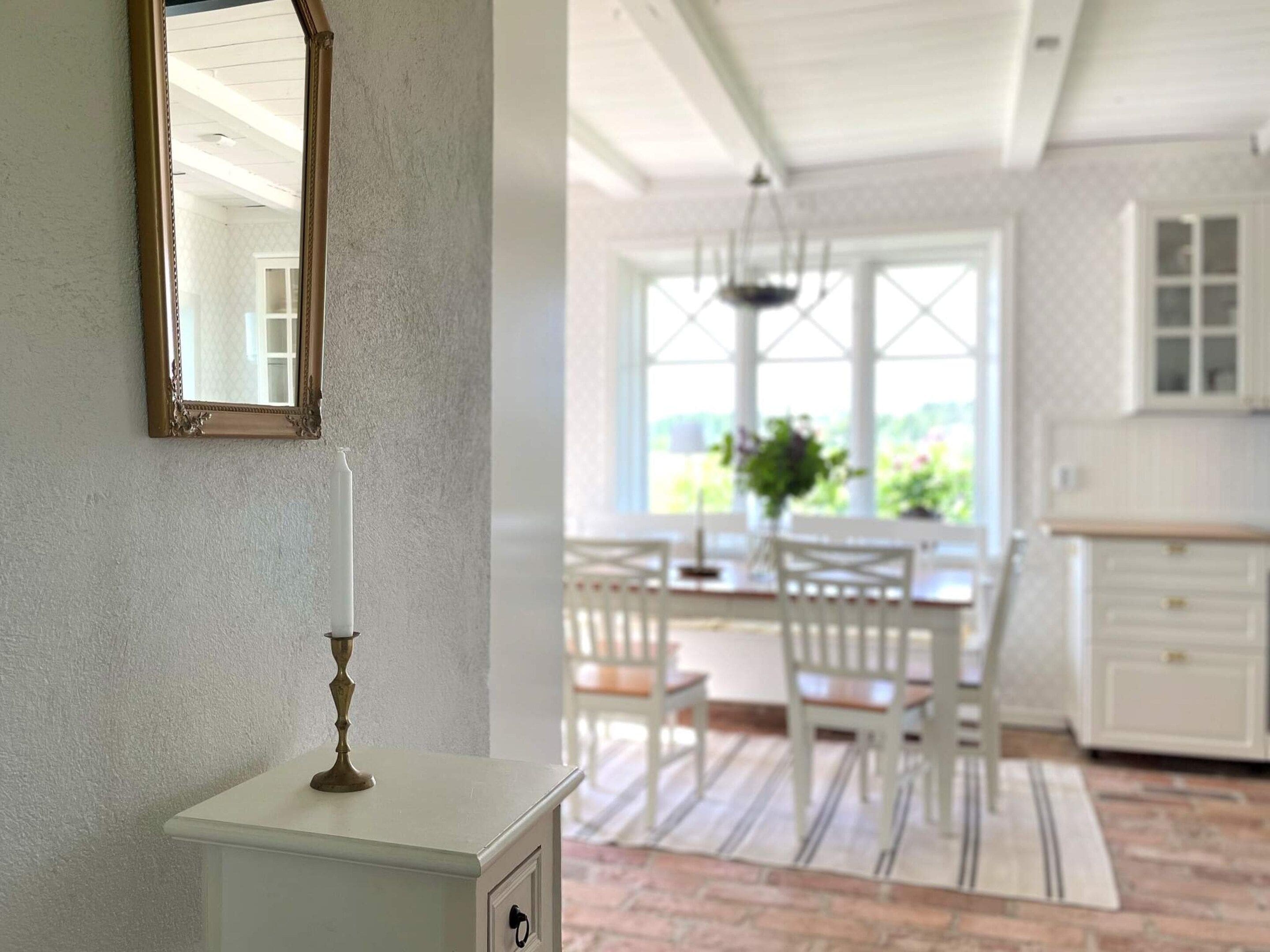 Une pièce comprenant une coiffeuse blanche, un miroir et un luminaire suspendu. On y trouve également un coin repas avec une table et des chaises, ainsi qu’une cuisine équipée de placards et dotée d’une fenêtre.
