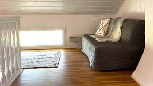 Une pièce avec un plancher en bois, un canapé gris avec une couverture, une rambarde blanche et une fenêtre avec des stores.