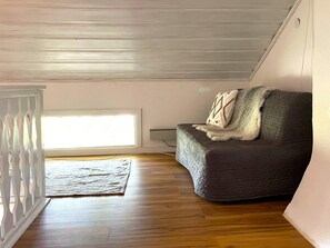 Une pièce avec un plancher en bois, un canapé gris avec une couverture, une rambarde blanche et une fenêtre avec des stores.