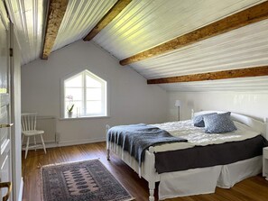 Un dormitorio con techo inclinado, un ventanal grande, una cama con colcha oscura, una silla blanca y una alfombra estampada.
