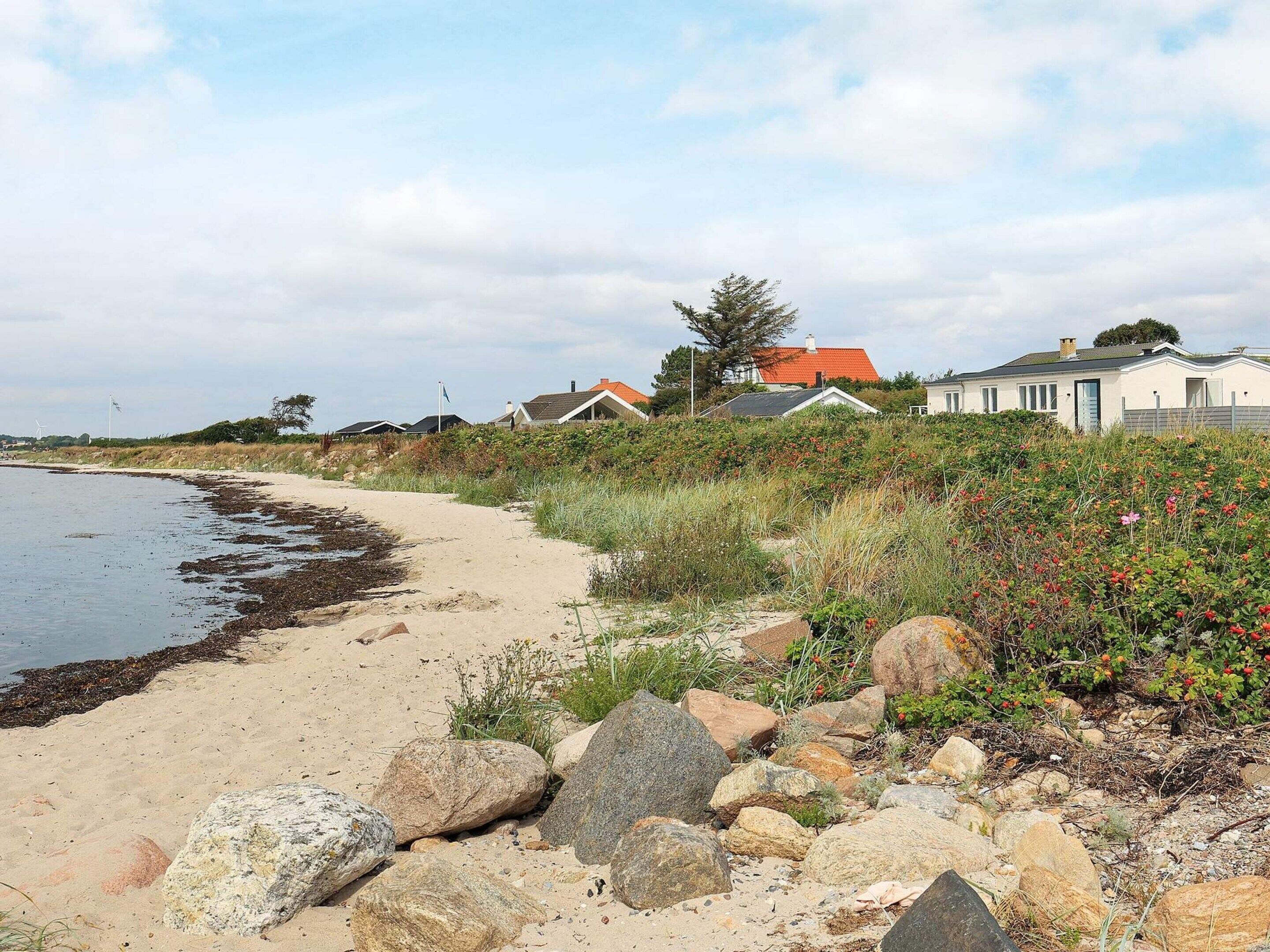 Une plage de sable avec des rochers et de la végétation, des maisons aux toits rouges et un ciel dégagé.