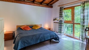 Un dormitorio con techo de madera, una cama con cubrecama azul acolchado, almohadas, una mesita de noche, una ventana con cortinas y vistas al verdor exterior.