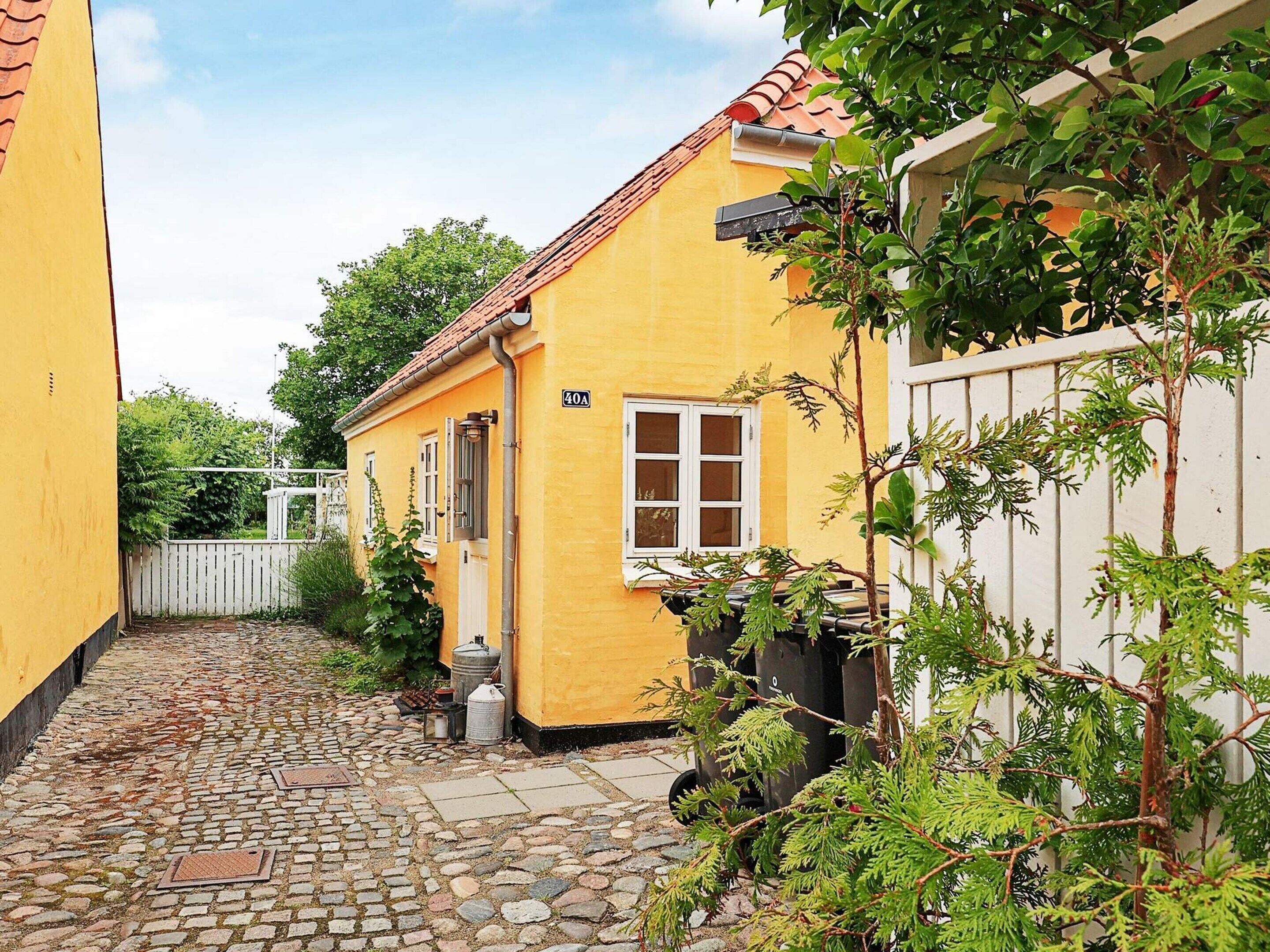 A yellow building with a cobblestone path, a white fence, and greenery.