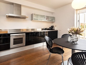 A modern kitchen with a built-in oven, a stainless steel range hood, and a dining area with a round table and black chairs. - 5 Star Holiday Home in Skagen (Skagen)