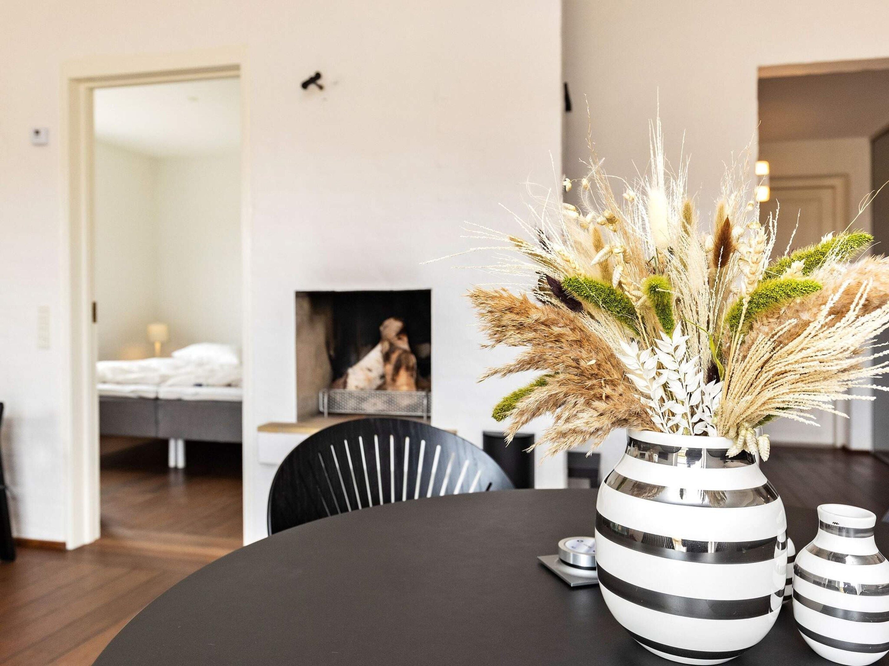 A modern living room with a dining table, a fireplace, and a vase with dried plants.