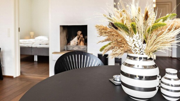 Un salon moderne avec une table ronde noire, un vase rayé contenant des plantes séchées et une cheminée en arrière-plan.