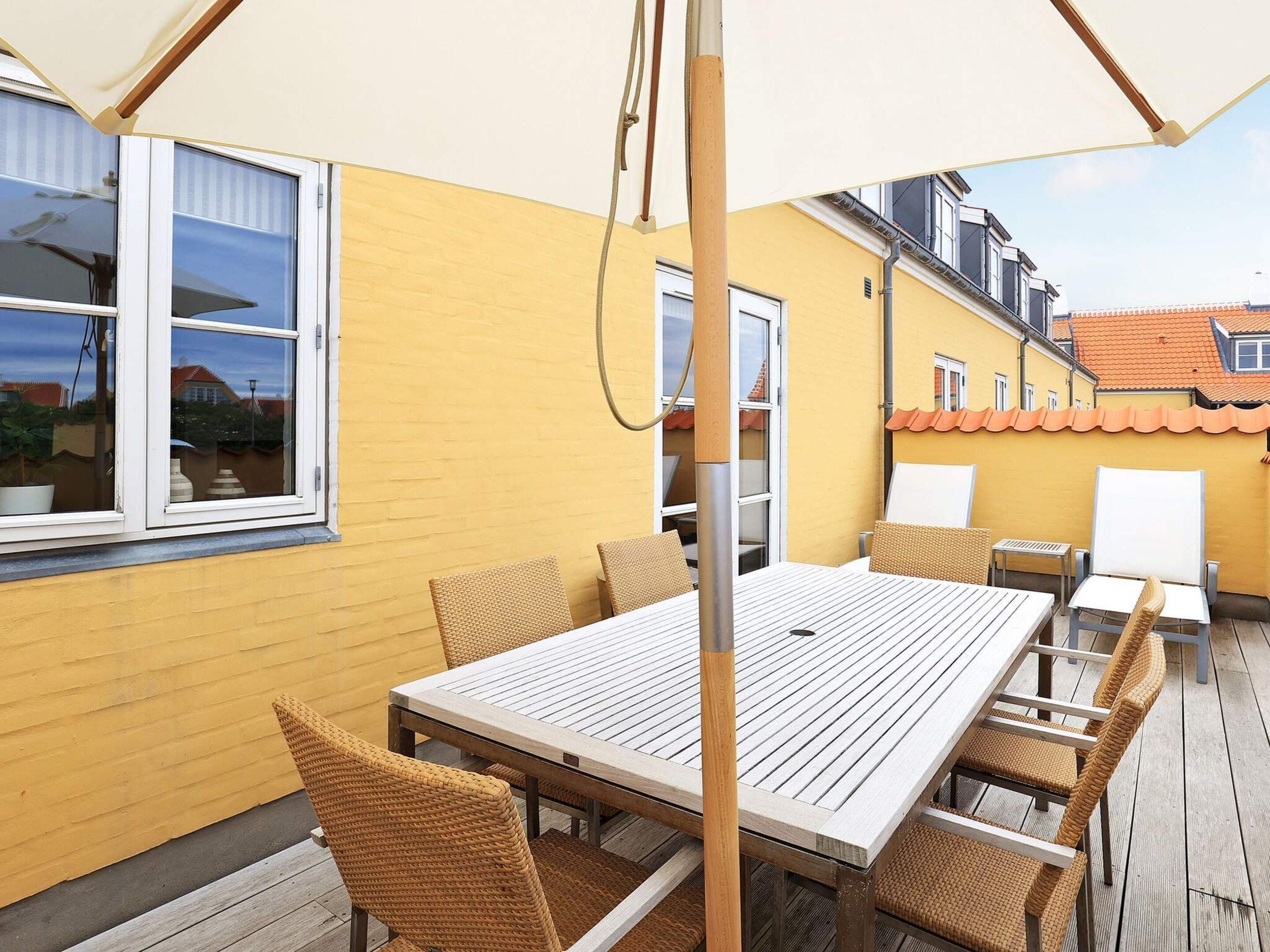 Une terrasse extérieure avec une table en bois et des chaises en osier, un parasol blanc et un mur jaune.