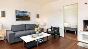 Un salon moderne avec un canapé gris, une table basse, un fauteuil noir, une cheminée et un lit à l’arrière-plan.
