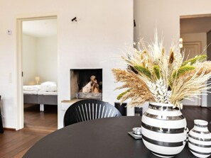 A modern living room with a round black table, a striped vase with dried plants, and a fireplace in the background.