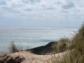 A coastal landscape with a sandy beach, grassy dunes, and a calm sea under a cloudy sky.