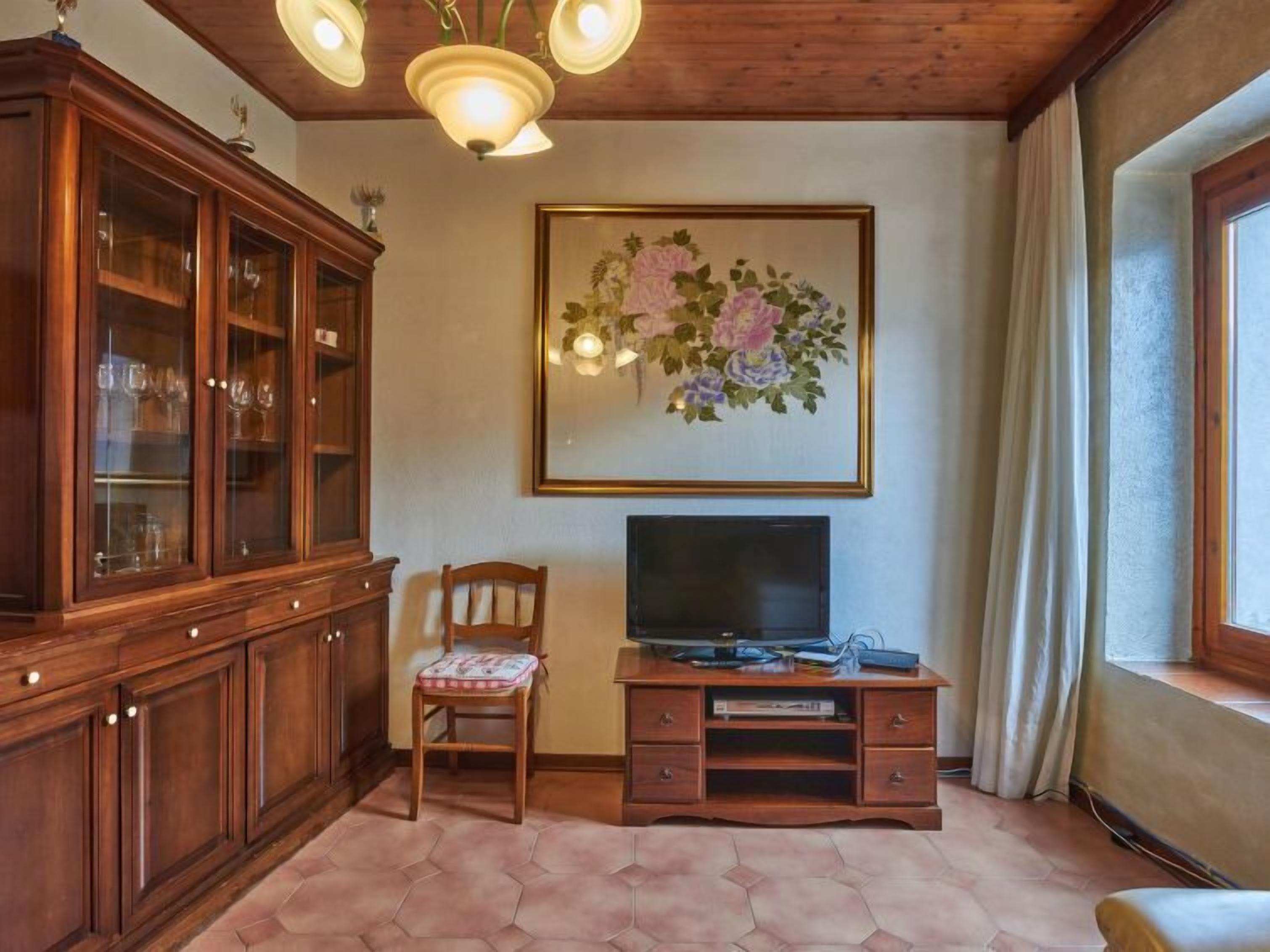 Ein Wohnzimmer mit einem Holzkabinett, einem Fernseher auf einem Holzständer, einem Blumenbild an der Wand und einem Fenster mit Vorhängen.