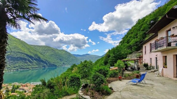 A scenic view of a lake with mountains in the background, a building with a balcony, and a patio area with a chair. - Casa Rosy by Interhome (Porlezza)