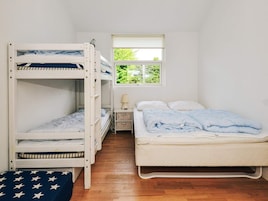 A room with bunk beds, a single bed, a small table with a lamp, and a window with a view of greenery.