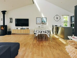 A modern living room with a fireplace, a dining area with a table, and a kitchen with a clock on the wall.