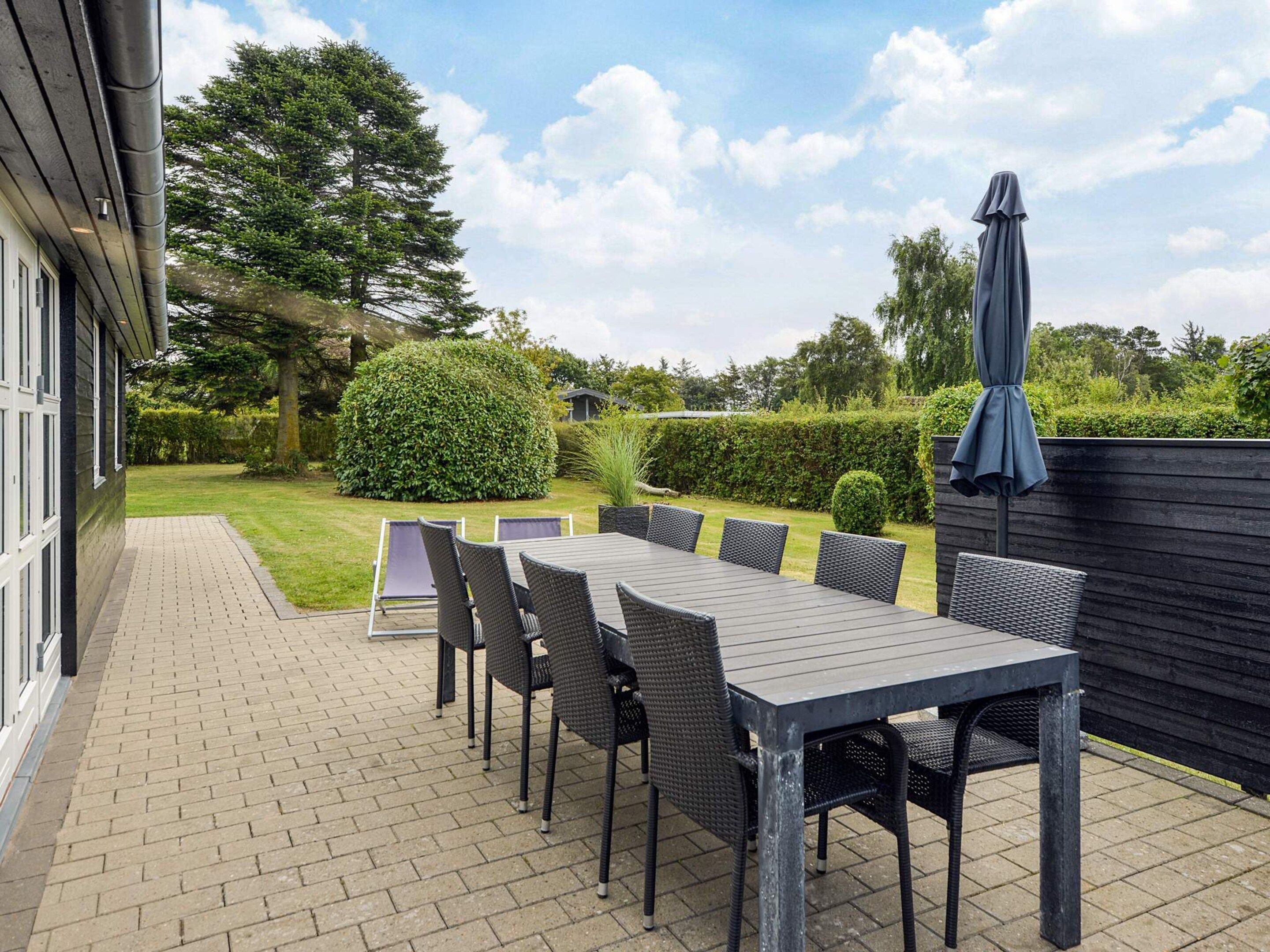 An outdoor patio with a long table and chairs, surrounded by a hedge and a wooden fence.