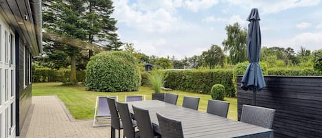 An outdoor patio with a long table and chairs, surrounded by a hedge and a wooden fence.