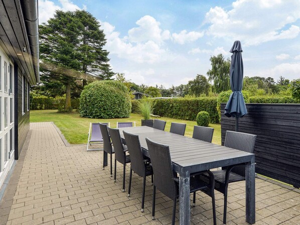 An outdoor patio with a long table and chairs, surrounded by a hedge and a wooden fence.