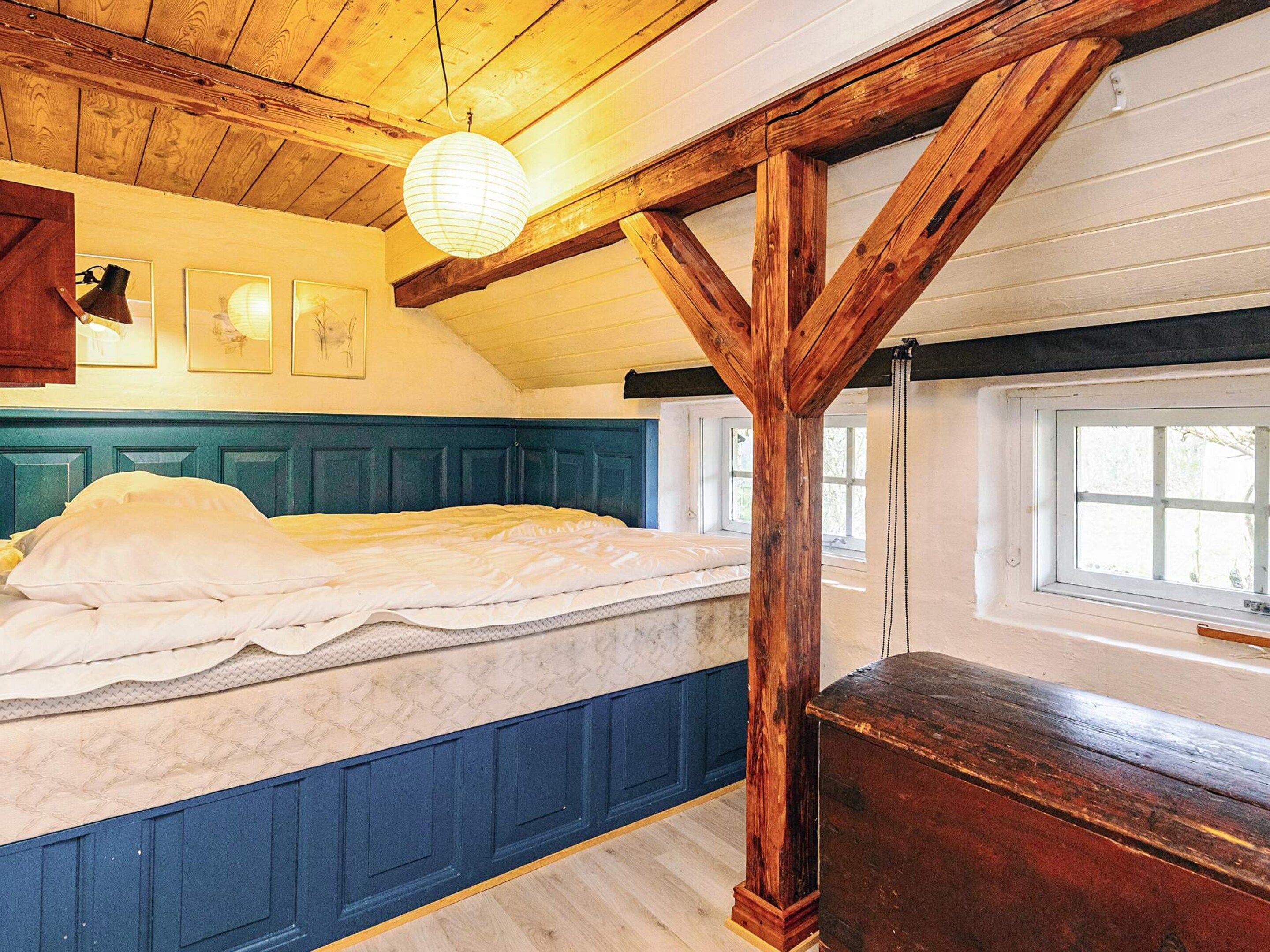 Quarto com cama, vigas de madeira, janela e luminárias fixadas na parede.