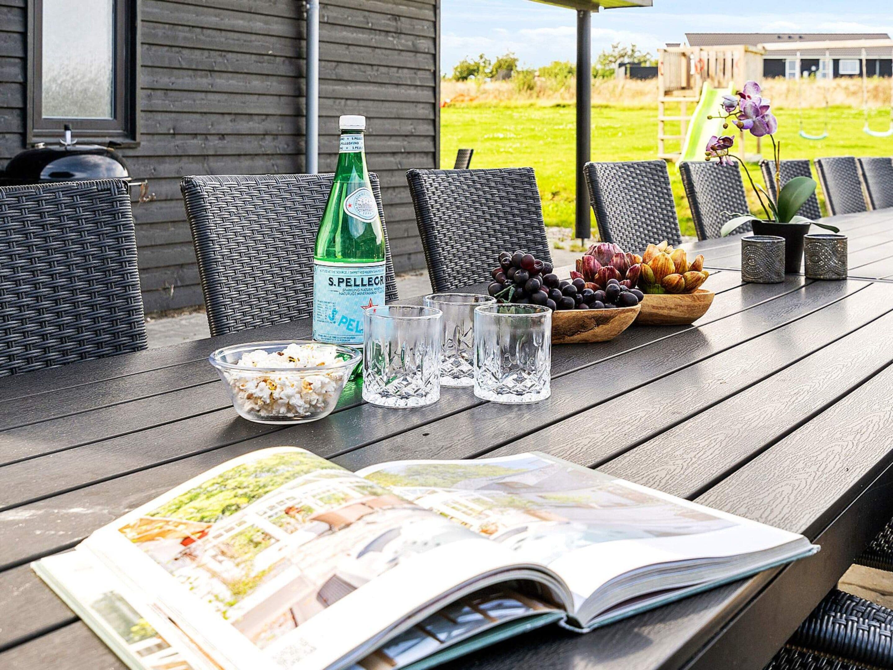 Una mesa de madera con una botella, vasos, un cuenco de fruta y una revista.
