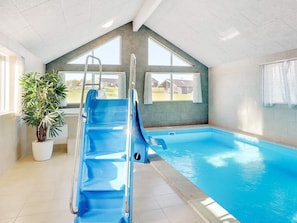 An indoor swimming pool with a blue slide, a potted plant, and a view of houses through large windows.