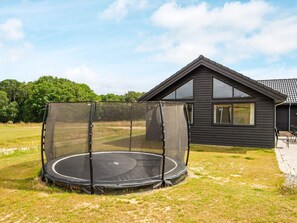 Un trampoline avec un filet de protection, installé sur une pelouse, une maison en bois sombre avec un toit en pente et un ciel dégagé.