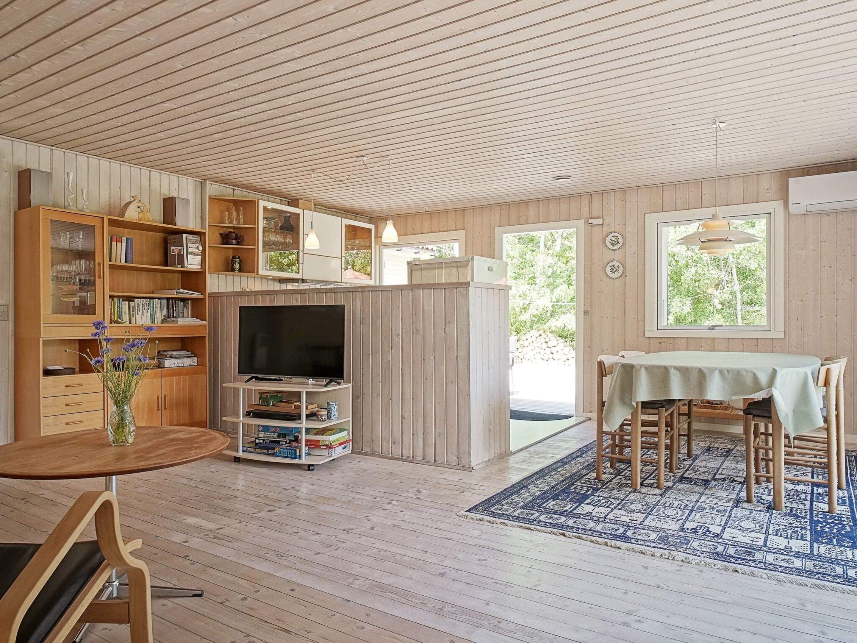 Ein gemütliches Zimmer mit Holzboden, einem Esstisch mit Stühlen, einem Fernseher auf einem Schrank und einem Bücherregal voller Bücher und Dekoration.
