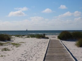 Un paseo marítimo de madera que conduce a una playa con vegetación escasa y un velero a lo lejos.
