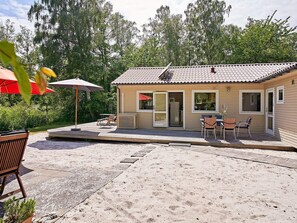 Um pequeno edifício térreo com telhado de telhas, um alpendre coberto com mobiliário de exterior e chão de areia.