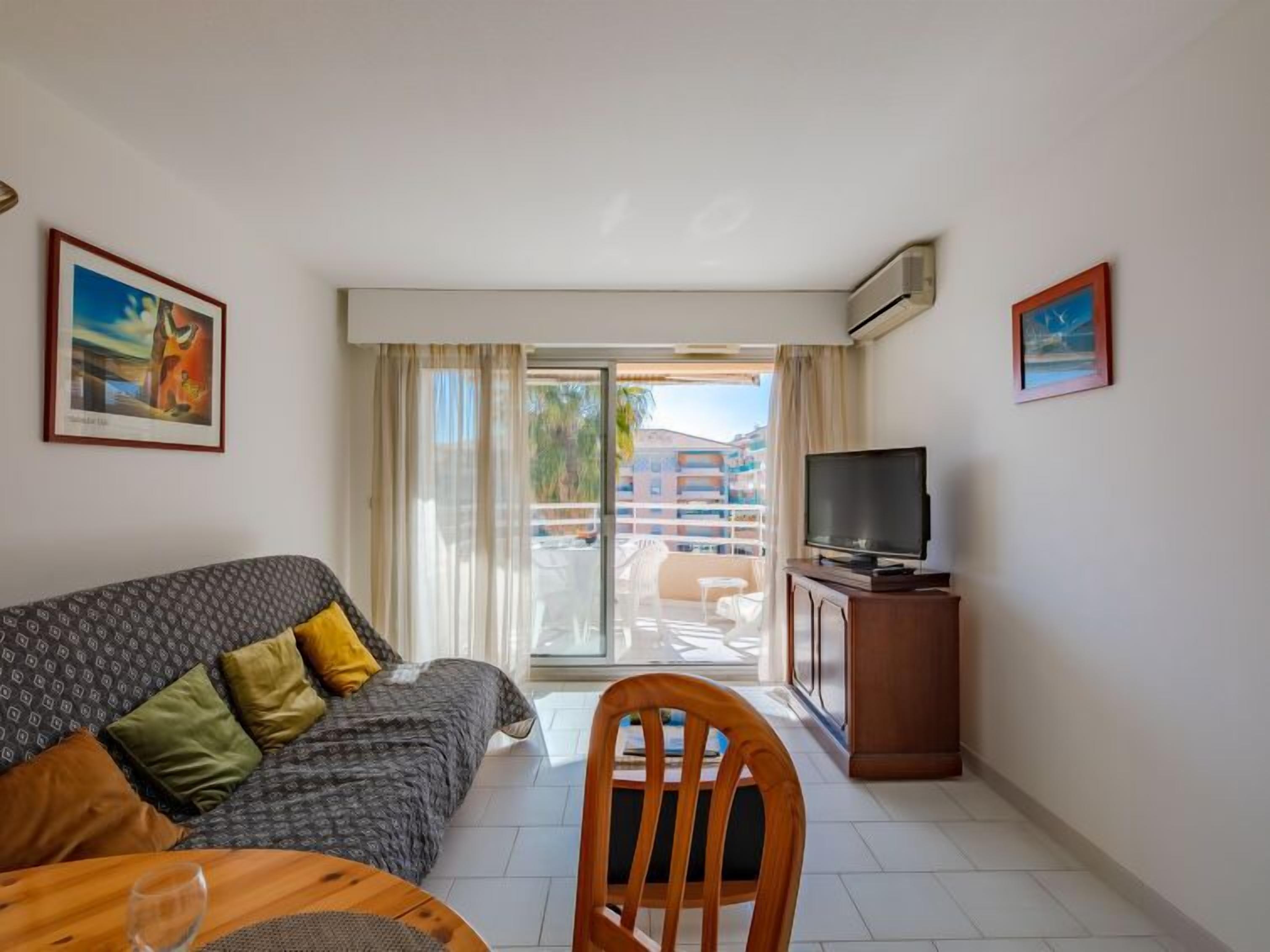 Ein Hotelzimmer mit einer Couch, einem Fernseher auf einer Holzkommode, einem Esstisch mit einem Stuhl und einem Balkon mit Blick auf Gebäude.