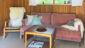 A cozy room with a wooden paneled wall, a pink sofa with cushions, a fluffy chair, and a small wooden table with a bowl. - 4 Person Holiday Home in Aakirkeby (Akirkeby)