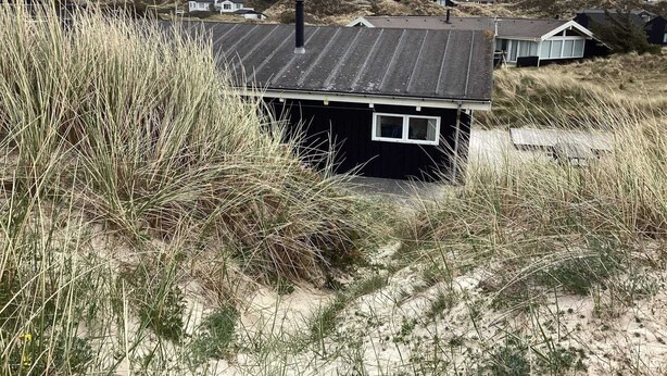A small, dark-colored house with a sloped roof, surrounded by tall grass and sand dunes.