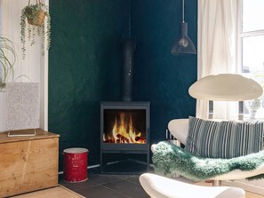 Una habitación con estufa de leña, una silla blanca con un plaid verde suave y una lámpara colgante.