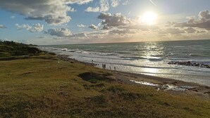 A coastal landscape with a grassy foreground, rocky shoreline, and a view of the ocean under a partly cloudy sky with the sun visible. - 8 Person Holiday Home in Gilleleje-by Traum (Gilleleje)