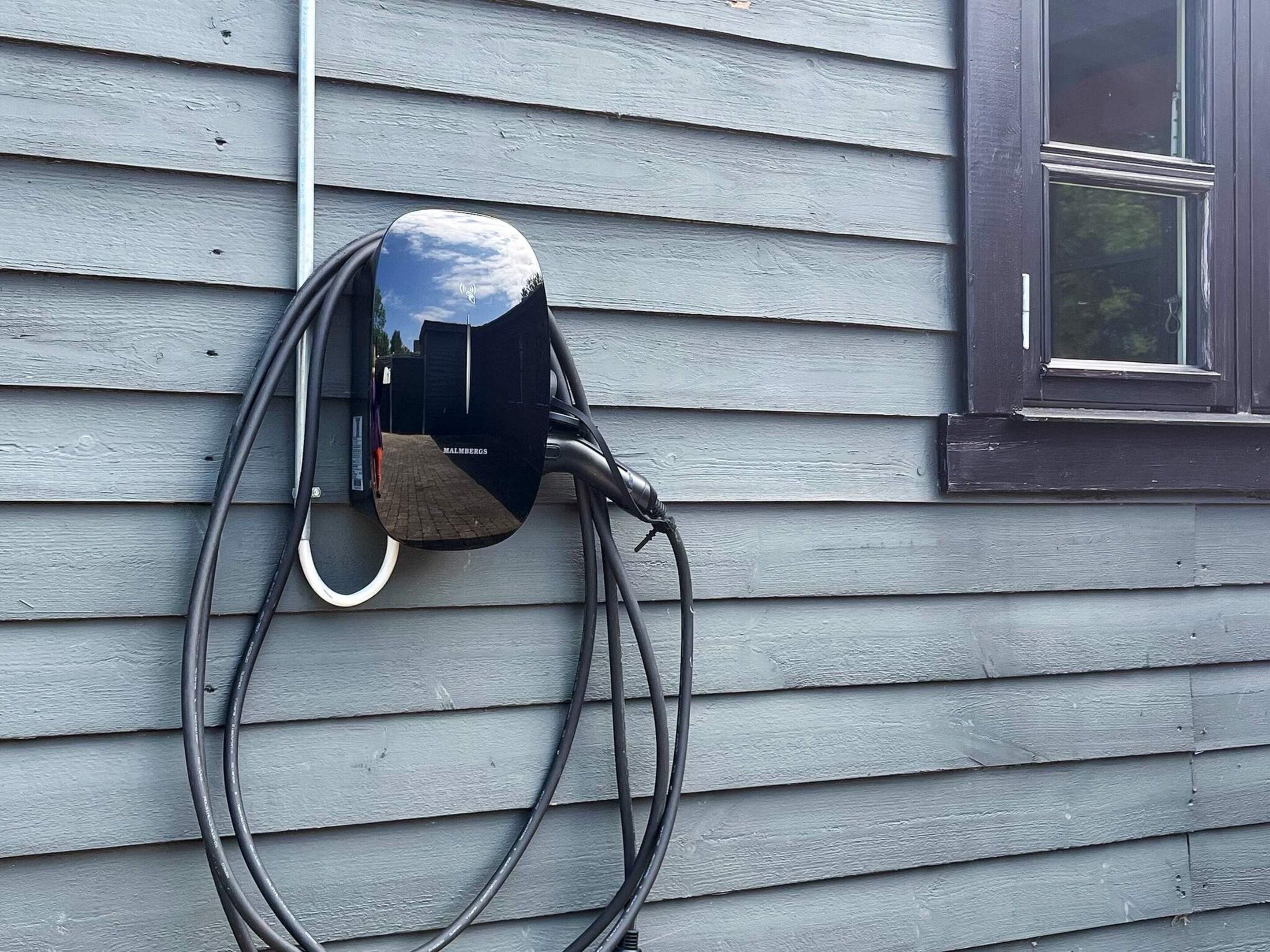Een zwarte laadpaal voor elektrische voertuigen, bevestigd aan een huis met horizontale beplating.