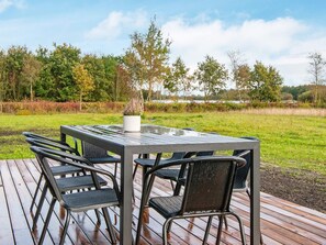 An outdoor dining area with a table and chairs, surrounded by a wooden deck and a view of trees and a body of water.