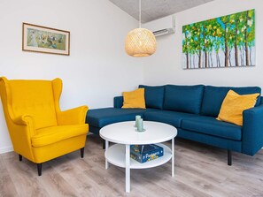 Una sala de estar con un sillón amarillo, un sofá azul, una mesa de centro blanca y un cuadro en la pared.