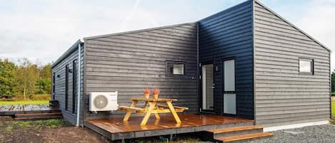 A modern, single-story building with a dark exterior, a wooden deck, and a large air conditioning unit.