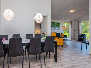 A modern dining area with a long table and chairs, a sofa, and a view of a garden through large windows.