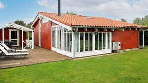 Een rode houten woning met pannendak, een overdekte patio met tafel en stoelen, en een groot raam.