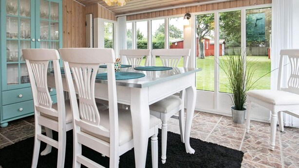 Een eetgedeelte met een witte tafel en stoelen, een hanglamp en uitzicht op de tuin door grote ramen.