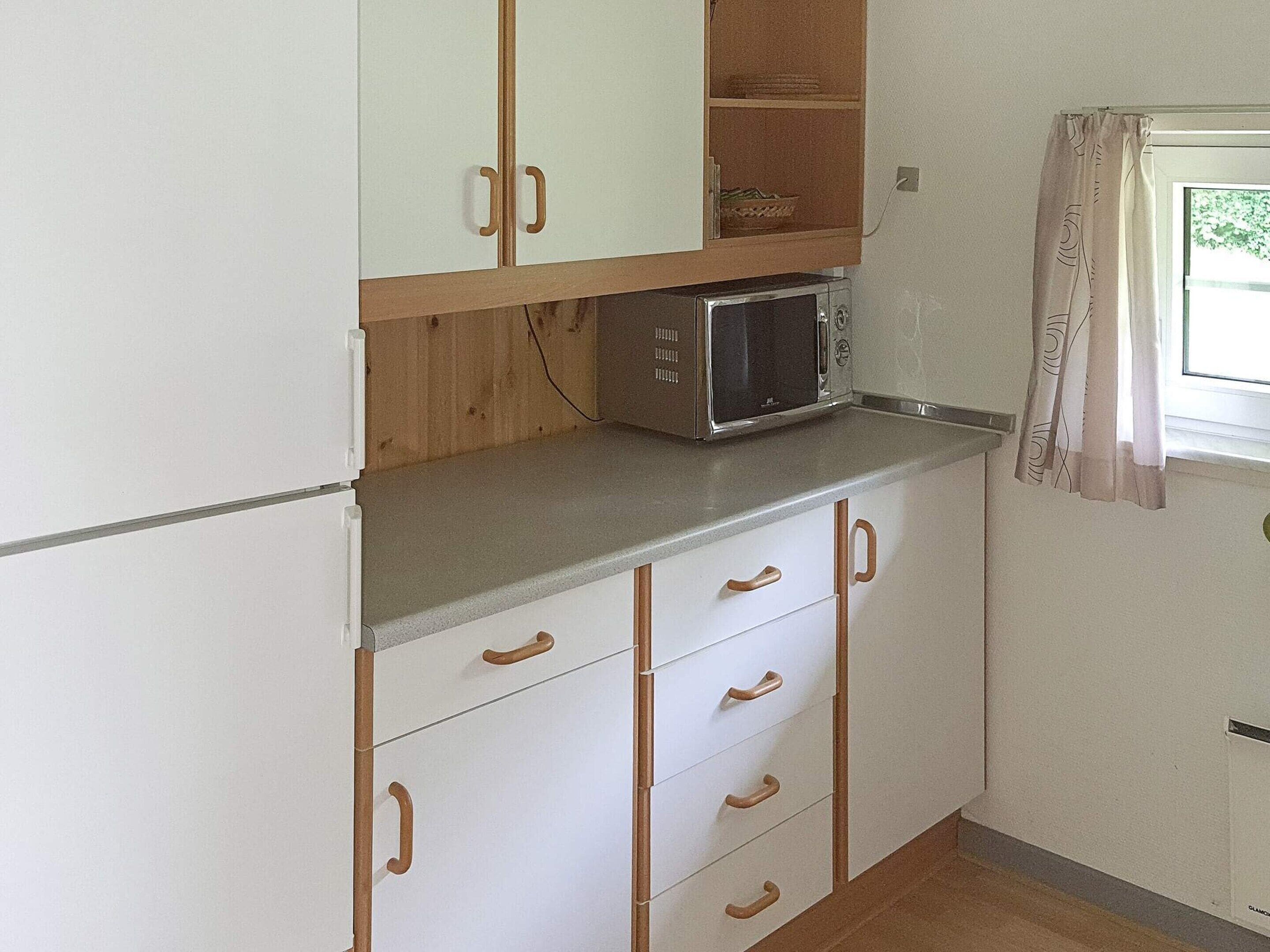 Eine kompakte Küche mit weißen Schränken, einer Mikrowelle und einem Fenster mit Vorhängen.