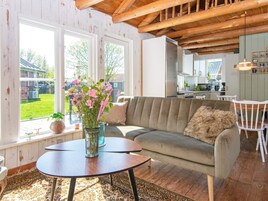 Un salón acogedor con un sofá, una mesita de centro y un jarrón con flores. Se ve un jardín y una casa a través de la ventana.