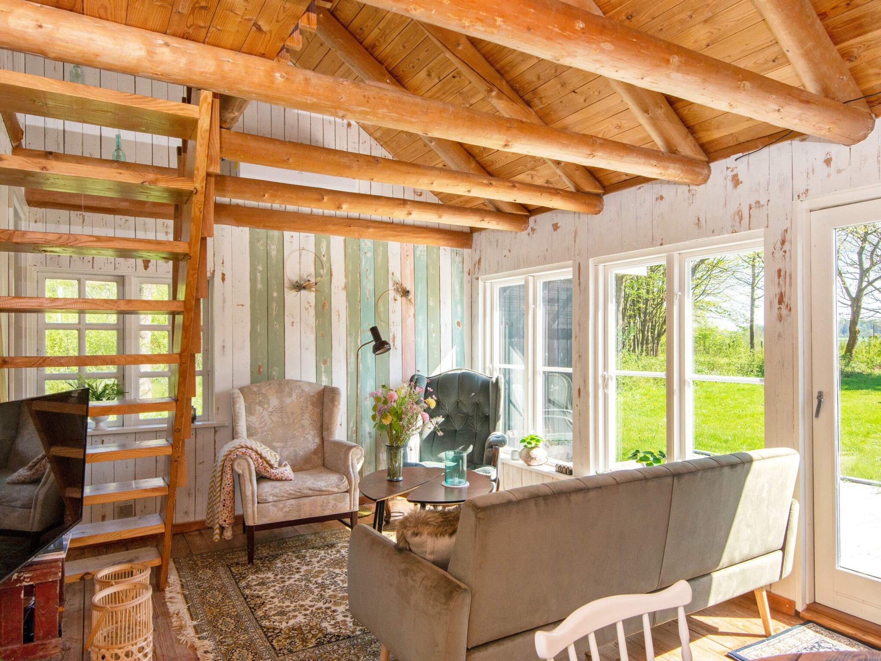 A cozy room with a wooden ceiling, a sofa, a chair, a TV, and a large window with a view of greenery.