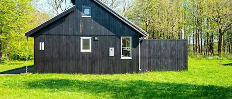 A black wooden building with a sloped roof, a small window, and a door, surrounded by a grassy field and trees.
