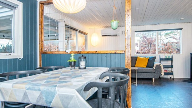 Un coin repas avec une table dressée pour six personnes, entourée de chaises. Un luminaire est suspendu au-dessus de la table et une fenêtre offre une vue sur les arbres.
