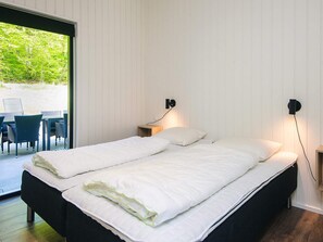 A double bed with white bedding, a bedside table with a lamp, and a view of outdoor chairs and greenery through a sliding door. - 5 Star Holiday Home in Sjolund (Sjølund)