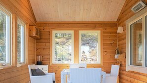 Une cabane en bois dotée d’un coin repas avec des meubles en osier blanc, de grandes fenêtres et une vue sur la forêt.