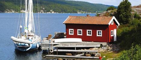 A red house by a dock with a sailboat, surrounded by greenery and a forested hill in the background.
