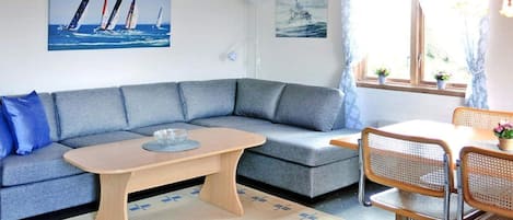 A cozy room with a grey sofa, a wooden coffee table, and a dining area with a table and chairs. There are two paintings on the wall, one of sailboats and one of a ship.