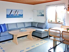 A cozy room with a grey sofa, a wooden coffee table, and a dining area with a table and chairs. There are two paintings on the wall, one of sailboats and one of a ship.